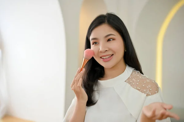 Attractive and beautiful young Asian woman applying makeup, applying brush-on on her cheek by makeup brush. Beauty and fashion concept