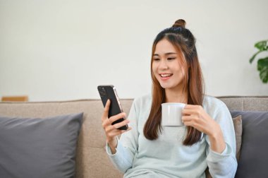 Akıllı telefonunu kullanan, sosyal medyasını karıştıran, sabah kahvesini yudumlayan ve oturma odasında dinlenen güzel Asyalı genç bir kadın..