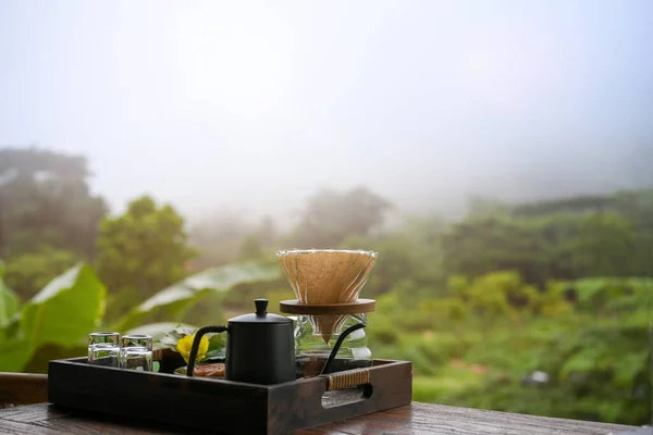 Pour-over coffee set in a wooden tray on wooden table over blurred beautiful green mountain forrest landscape view. Driper, kettle, coffee beans. close-up image