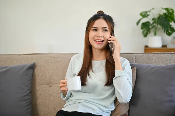 Attractive young Asian female on the phone with her friend, sipping morning coffee while relaxing on her sofa in the living room.