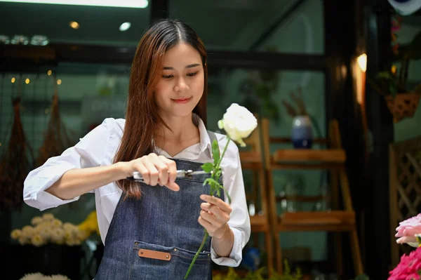Beautiful and cheerful young Asian female florist or flower shop owner making a beautiful roses bouquet, cutting a rose stalk by scissors.