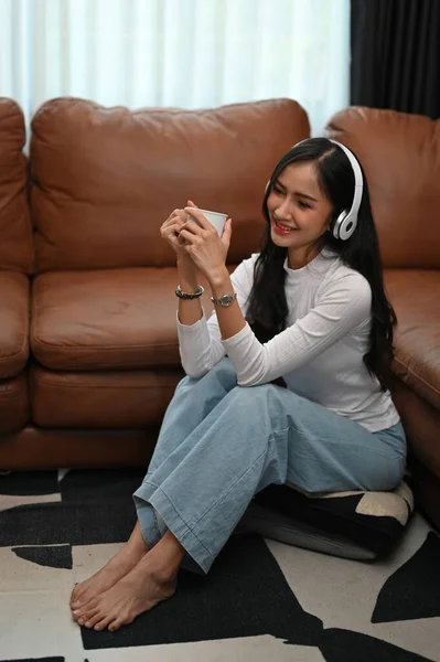肖像画 陽気で幸せな若いアジアの女性は 彼女のヘッドフォンを介して音楽を聴く コーヒーをすすり その後リビングルームの床に座って — ストック写真