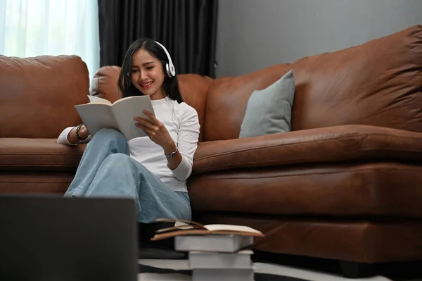 漂亮而轻松的年轻亚洲女性喜欢在现代的客厅里一边通过耳机听音乐 一边读她最喜欢的书 — 图库照片