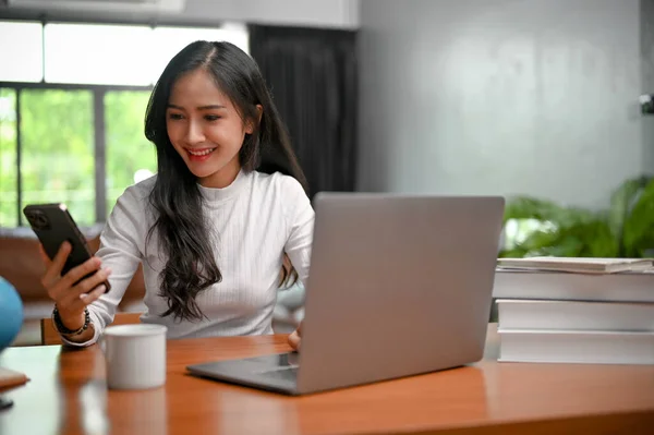 Pretty and charming young Asian female using her smartphone at her working desk. chatting, online shopping, scrolling on phone, downloading application, sending email, reading web blog.
