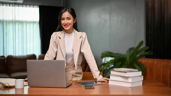 Confident and successful millennial Asian businesswoman or female executive manager stands at her desk in her modern vintage private office.