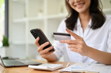 Ofis masasında elinde cep telefonu ve kredi kartı olan mutlu Asyalı genç bir iş kadını faturalarını ödemek için mobil bankacılık başvurusunu kullanıyor. Çevrimiçi ödeme kavramı