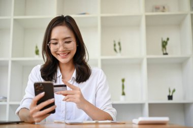 Gülümseyen ve çekici gözlüklü Asyalı kadın kredi kartıyla online alışveriş yapıyor, mobil bankacılık uygulaması kullanıyor..