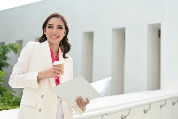 Gorgeous millennial Asian businesswoman or female business consultant holding a document folder and a coffee cup, smiling and looking at the camera, standing outside of the building.