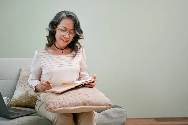 Happy and peaceful 60s retired asian woman in glasses reading a book while relaxing in her home living room.