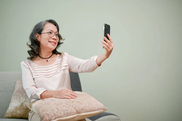 Cheerful and beautiful 60s retired Asian woman in glasses, using her smartphone to take a picture of her face, taking selfie while relaxing in her living room.