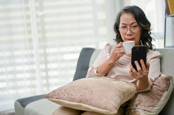 Concentrated 60s retired asian woman wearing glasses using her smartphone to read an online news while sipping coffee in the living room.