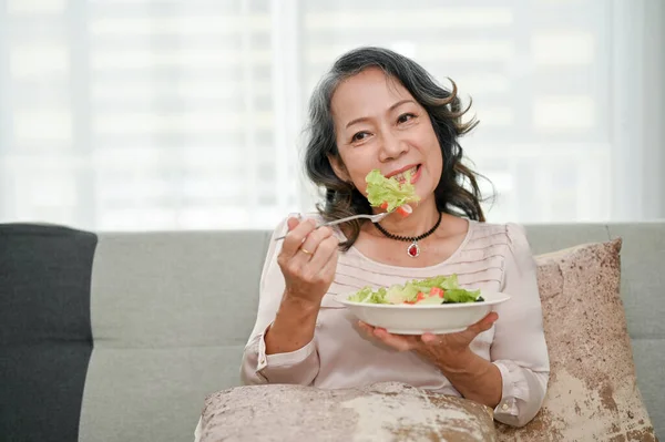 迷人的60多岁的退休亚洲女人喜欢在客厅休息的同时吃健康的绿色沙拉 健康生活方式概念 — 图库照片