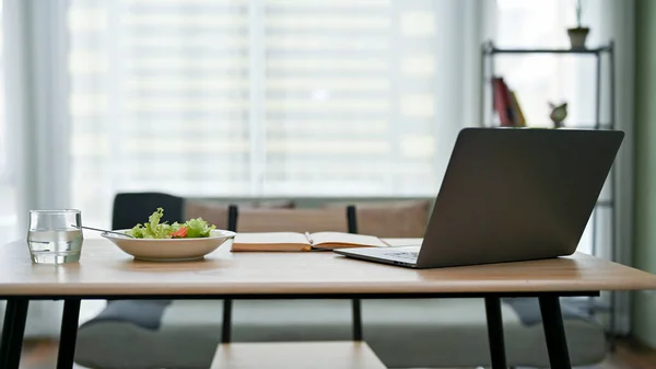 Modern urban office workspace with laptop computer, book, a glass of water and a salad bowl on the table over blurred background of sofa. Home office or private office interior