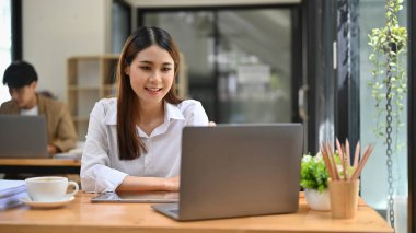 Çekici ve güzel Asyalı genç iş kadını ya da kadın ofis çalışanı dizüstü bilgisayarına bakıyor, ofis bilgisayarındaki iş görevleri üzerinde çalışıyor..