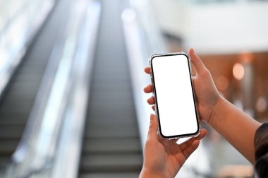 Beyaz ekranlı akıllı telefon modeli arka plandaki bulanık alışveriş merkezinin yürüyen merdiveninin üzerinde bir kadının elinde. yakın plan resim