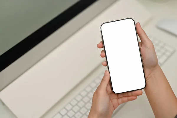 A smartphone white screen mockup is in a woman hands over blurred minimal white office desk in the background. close-up image