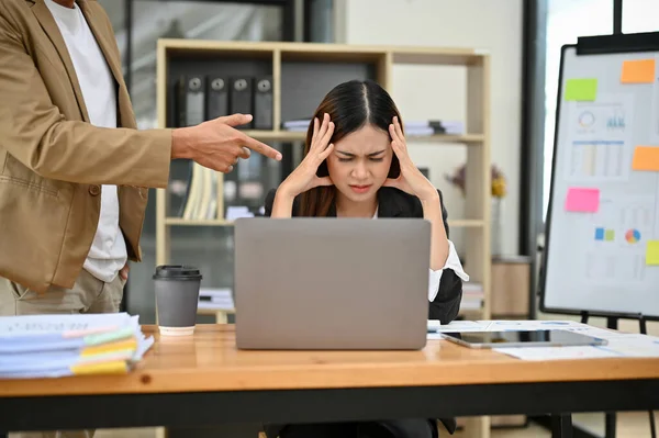 Stressed young Asian female office employee getting complaint by her boss, feeling tired of work. Unhappy and mad male boss yelling and pointing finger at his female employee.