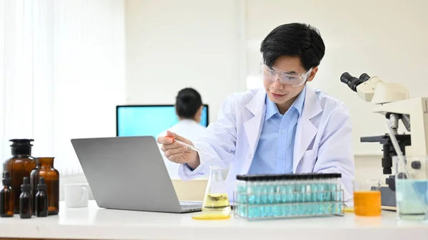 Smart young Asian male scientist or chemist in goggles and gown adjusting a liquid sample in a conical beaker, working in the lab office.