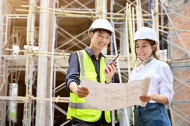 Asyalı profesyonel bir erkek mühendis ve çekici bir Asyalı kadın mimar inşaat alanının önünde bir plan arıyor ve tutuyor..