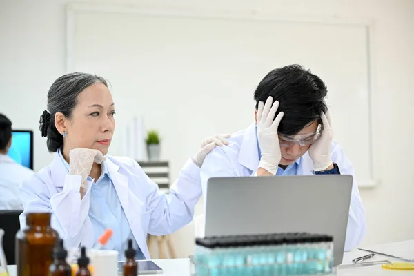 Stressed young Asian male scientist upset and dissatisfied with his medical experiment project while working in the lab with senior female professor.