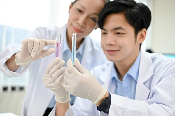 Happy and cheerful young Asian male scientist and senior female supervisor looking at the test tubes together. Pharmaceutical concept