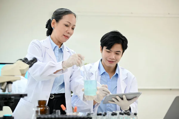 Professional Asian-aged female scientist adjusts a new chemical liquid in a beaker while a young male scientist records the chemical experiment results on a tablet.