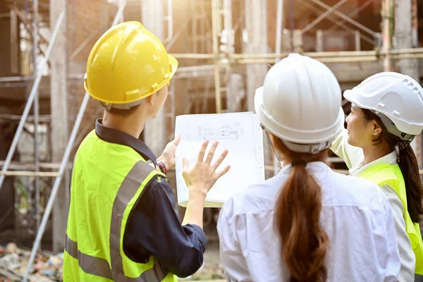 A professional and smart Asian male engineer explaining the building plan with a blueprint to a female architect and a businesswoman at the construction site.