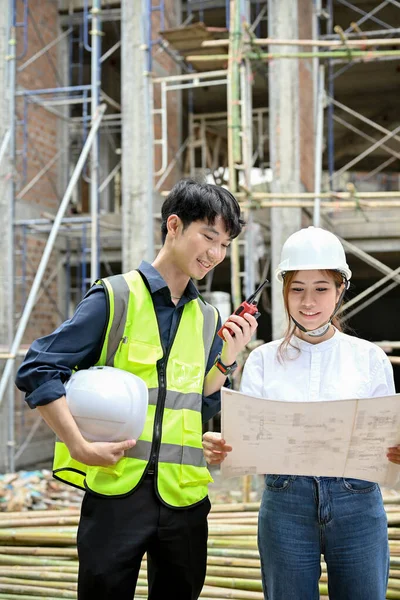 Smart and handsome young asian male engineer and beautiful female architect in the construction site, looking and checking the building blueprint together.