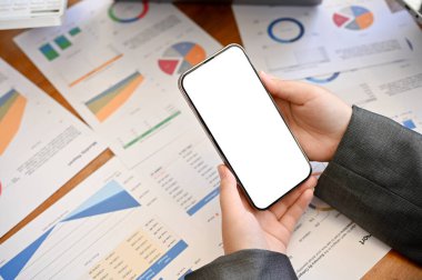 Ofis masasının üzerinde elinde akıllı telefon beyaz ekran maketi tutan bir iş kadını elleri ve mali rapor belgeleri. yukarıdaki görünüm