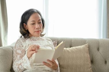 Çekici bir Asyalı kadın rahat oturma odasında kitap ya da roman okumaya odaklanıyor..