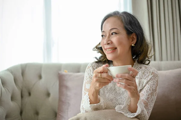 美しいアジア系の女性は 彼女の居心地の良いリビングルームで朝のコーヒーを飲みながらリラックスし 窓の外を眺め 幸せを夢見て — ストック写真