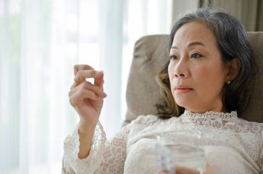 Stresli ve bunalımlı Asyalı orta yaşlı bir kadın oturma odasında ilaç ya da hap alıyor. Sağlık sorunları kavramı.