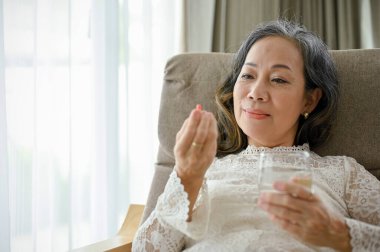 Mutlu ve çekici Asyalı orta yaşlı kadın oturma odasında dinlenirken ilaç ya da vitamin alıyor. Sağlık hizmeti kavramı