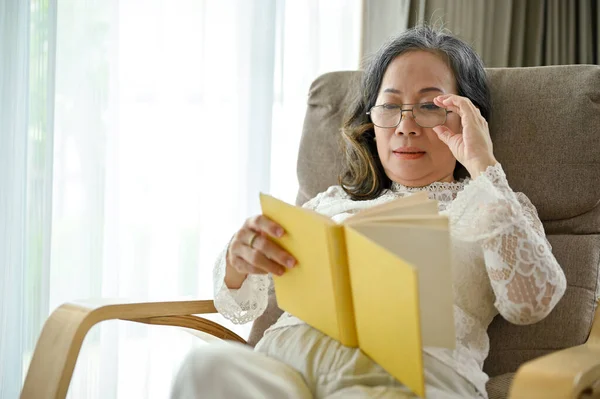 眼鏡をかけたリラックスしたアジア系の中年女性が彼女のリビングルームで本を読んで快適なアームチェアに座っています — ストック写真