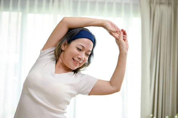 Healthy and happy Asian-aged woman exercising in her home living room, practicing yoga, stretching her arms. Healthy and wellbeing lifestyle.