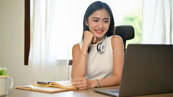 迷人迷人的年轻亚洲女商人或女企业家坐在自己的办公桌前 对她的商业项目进行思考和规划 手牵着下巴 微笑着看着她的笔记本电脑屏幕 — 图库照片