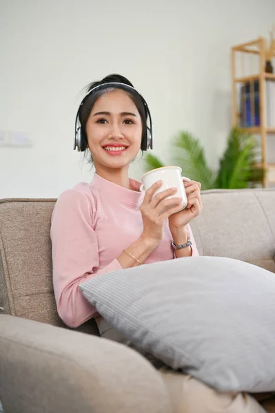 커피를 마시고 소파에서 쉬면서 헤드폰으로 음악을 아름다운 아시아 소녀의 — 스톡 사진