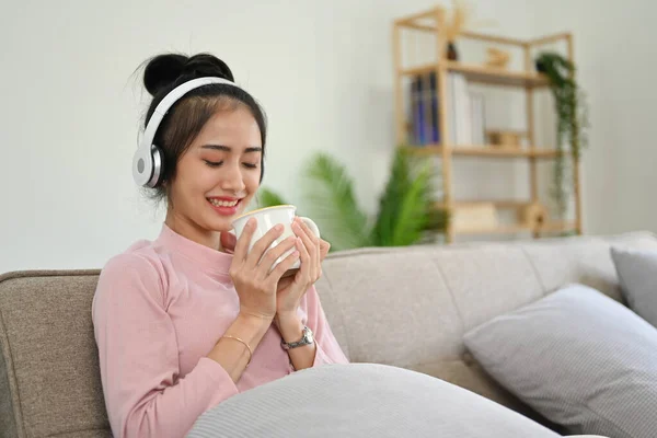 Calm and peaceful young Asian female in casual clothes listening to music through her wireless headphones while smelling her coffee and relaxing on sofa.