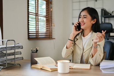 Çekici ve çekici Asyalı iş kadını ofis masasında otururken iş arkadaşıyla telefonda konuşmaktan hoşlanıyor..