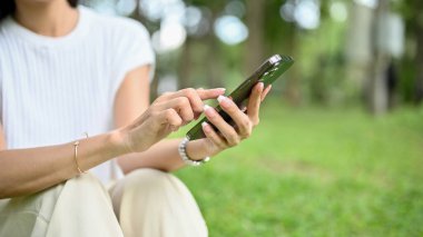Genç Asyalı bir bayanın parkta dinlenirken cep telefonuyla sohbet etmesi ya da online makaleler okuması. kırpılmış resim