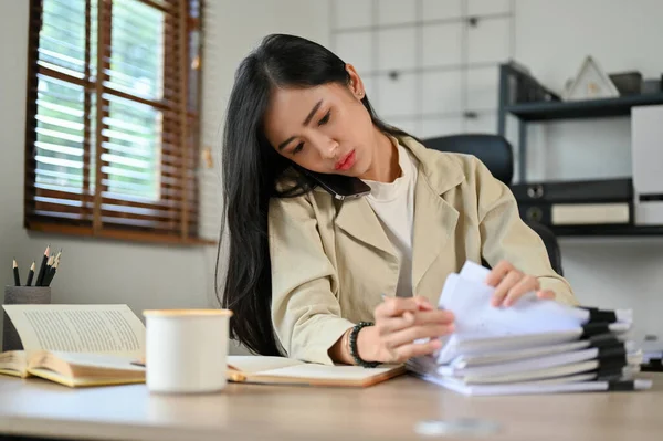 Professional and busy Asian businesswoman or female accountant focused on talking with her client on the phone while working on her documents at desk.