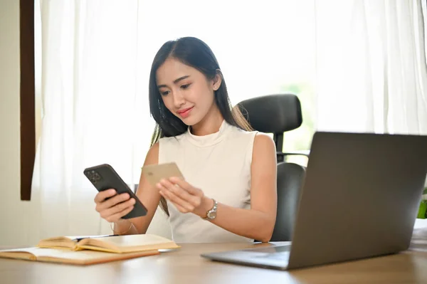 迷人的年轻亚洲女商人坐在写字台前 用她的在线移动银行申请在购物网站上登记她的信用卡 — 图库照片