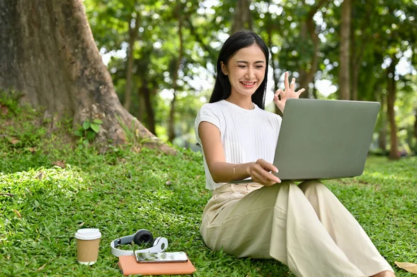漂亮的年轻亚洲女大学生或自由职业者在城市公园放松的时候 手签不错 在网上开会 或在笔记本电脑上打视频电话 — 图库照片