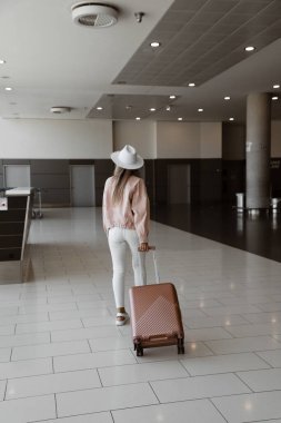 Genç kız kalkıştan önce havaalanında duruyor, beyaz pantolon, pembe ceket ve şapka giymiş bir model, elinde pembe bavul, geriye doğru giden bir kadın.
