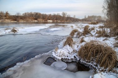 Unfrozen river in winter. Chunks of ice float in the water near the shore. Nearby islets with yellow, dry grass.