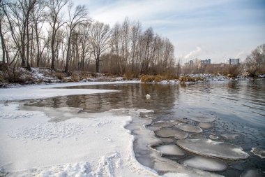 Unfrozen river in winter. Chunks of ice float in the water near the shore. In the distance there is a forest and an industrial plant.