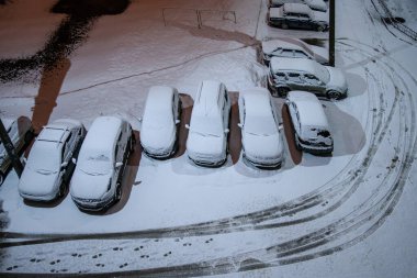 Karla kaplı araçlar geceleyin yüksek katlı bir binanın avlusuna park edilmiş. Fenerlerin ışığında kar kıvrılır. Karda yoldan geçenlerin ve arabaların izleri var..
