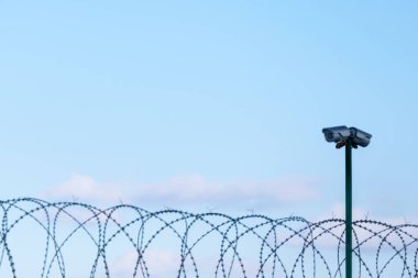 Dikenli tellerin üzerinden mavi bir gökyüzüne bakan güvenlik kameraları. Korunaklı bölge, giriş yasak. Yasadışı giriş kavramının korunması ve karşıtlığı