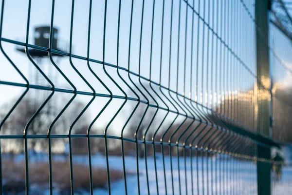 Border fence with barbed wire in front of the surveillance tower in a snow-covered field against a blue sky. Border protection and counteraction to illegal migration concept