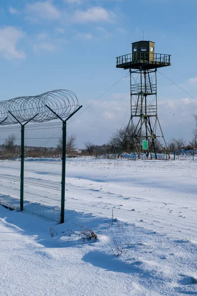 Border fence with barbed wire in front of the surveillance tower in a snow-covered field against a blue sky. Border protection and counteraction to illegal migration concept